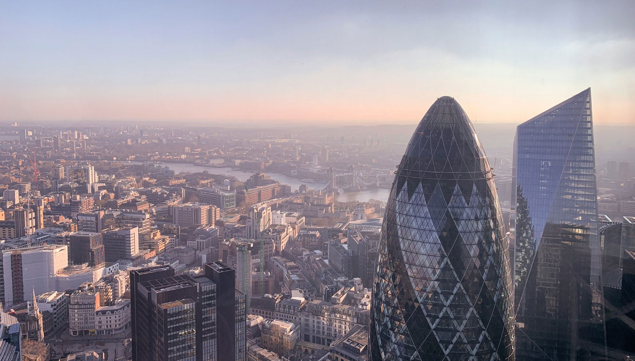 Aerial view over the City of London at dusk, with the Gherkin and the Scalpel in the foreground and the Thames beyond.
